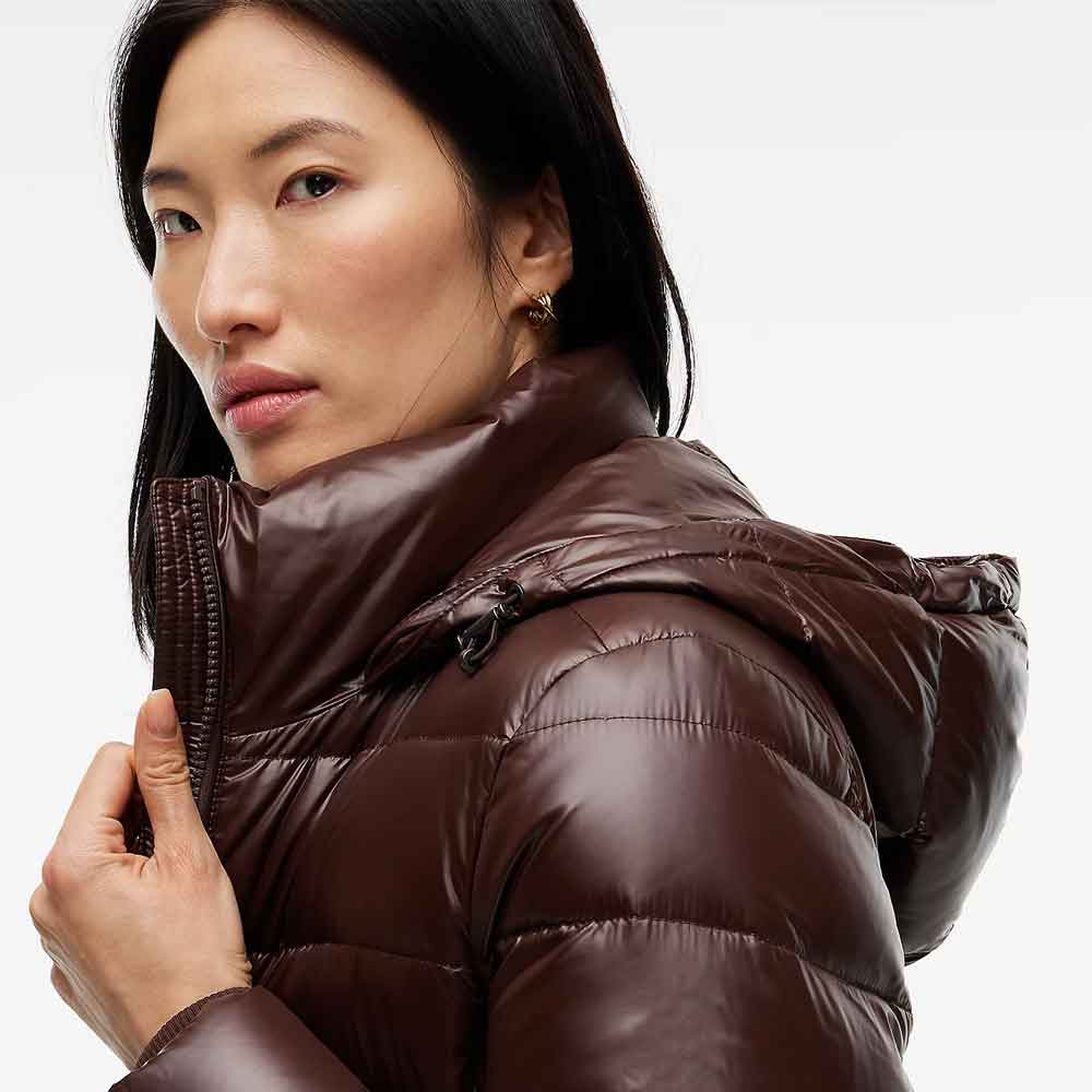 A close-up of a woman wearing a shiny dark brown puffer jacket with a high collar and hood, photographed against a clean light gray studio background.