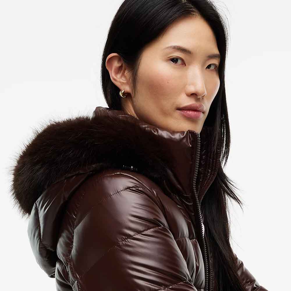 A close-up shot of a woman wearing a glossy dark brown puffer jacket with a thick dark brown fur-trimmed hood, set against a plain light gray background.