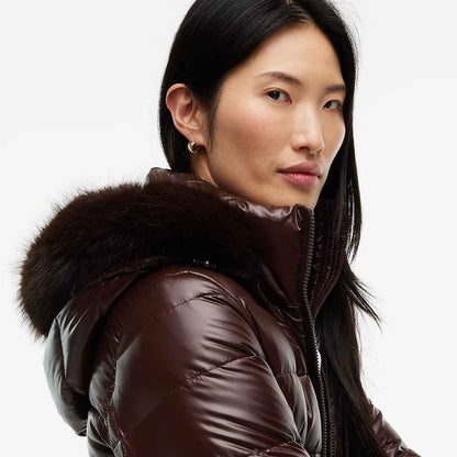 A close-up shot of a woman wearing a glossy dark brown puffer jacket with a thick dark brown fur-trimmed hood, set against a plain light gray background.