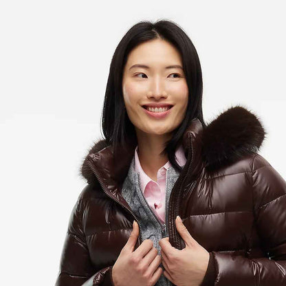 A woman wearing a dark brown puffer jacket with a fur-trimmed hood, layered over a pink shirt and gray sweater, photographed against a plain light gray background.