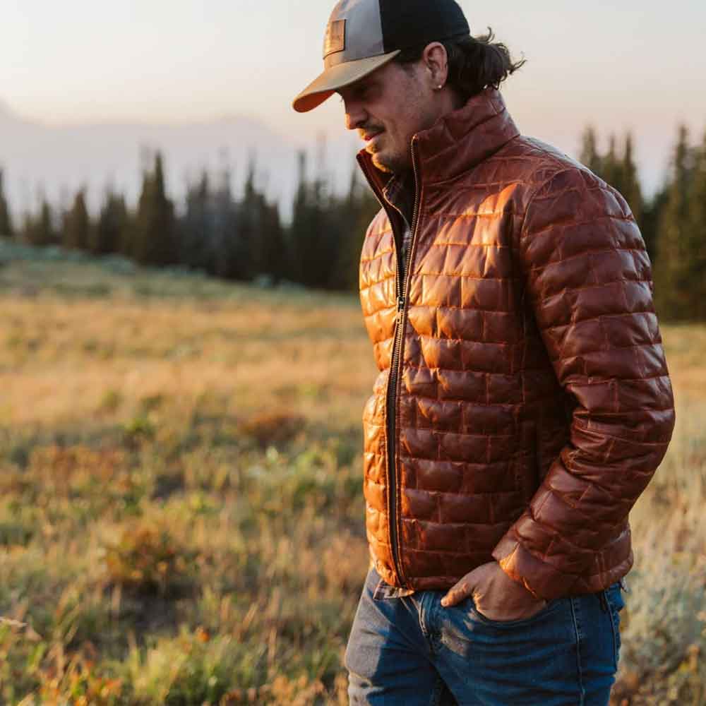 Man wearing a brown puffer jacket and cap in a field with trees in the background