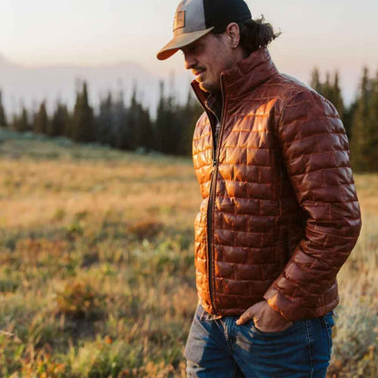 Man wearing a brown puffer jacket and cap in a field with trees in the background