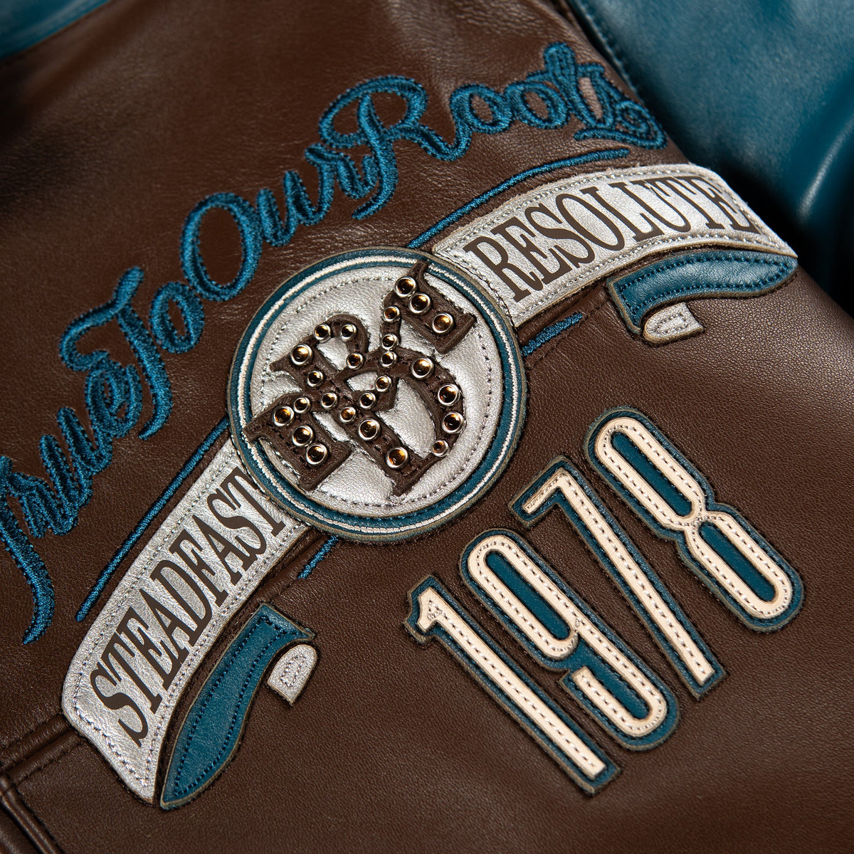 Close-up of a brown leather jacket featuring embroidered patches and text, including 'True To Our Roots', 'Steadfast Resolve', '1978', and a circular emblem with metallic studs.