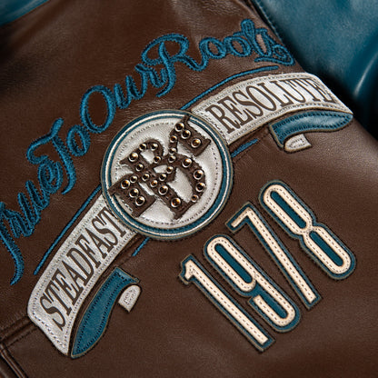 Close-up of a brown leather jacket featuring embroidered patches and text, including 'True To Our Roots', 'Steadfast Resolve', '1978', and a circular emblem with metallic studs.