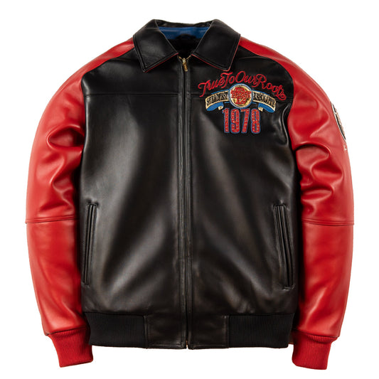 A black and red leather jacket with a full-zip front, ribbed collar, cuffs, and hem, featuring a 'True To Our Roots' embroidered patch on the left chest.